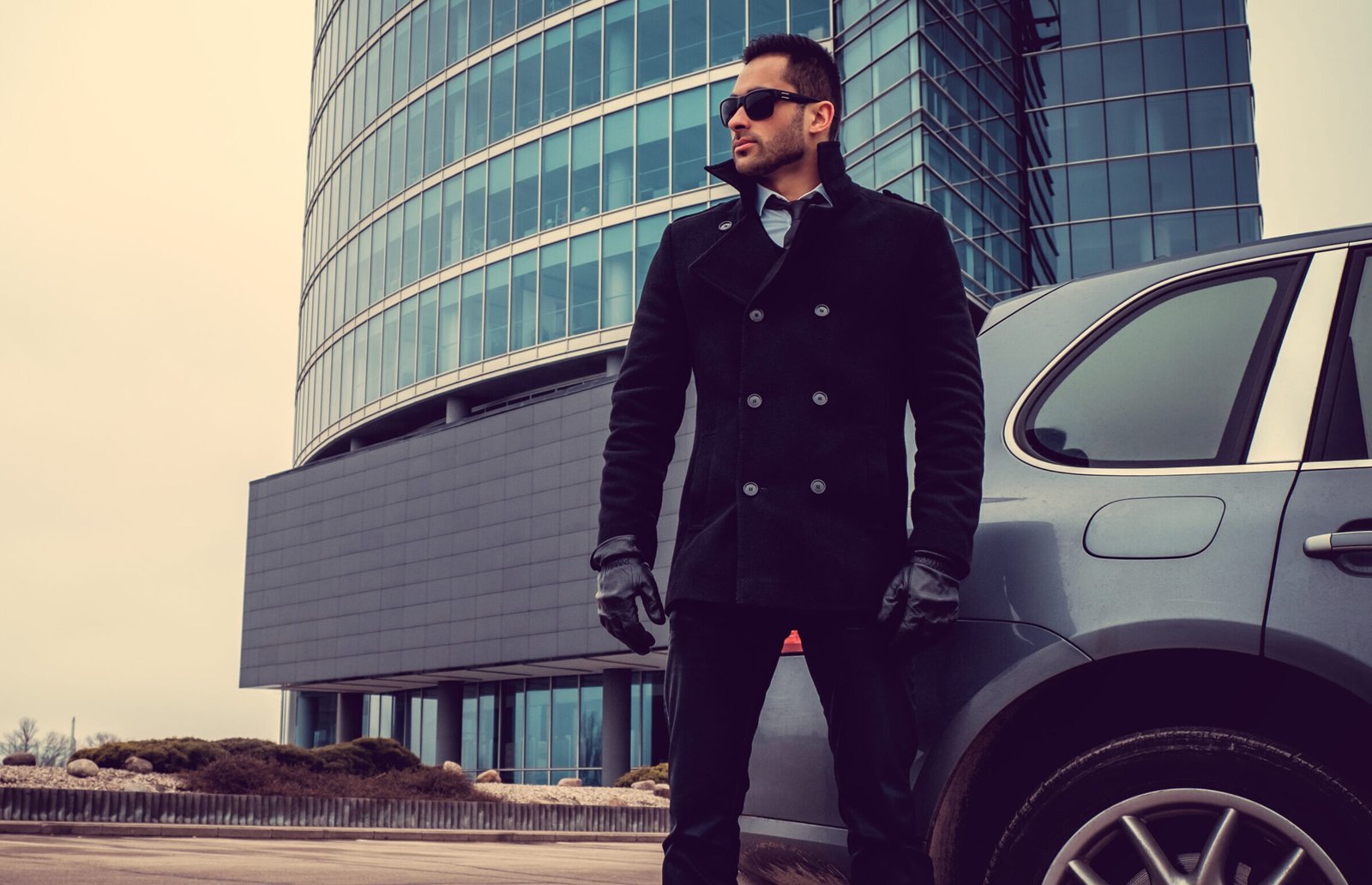 Stylish shirt hair male in sunglasses and warm autumn jacket posing near car in glass building background.